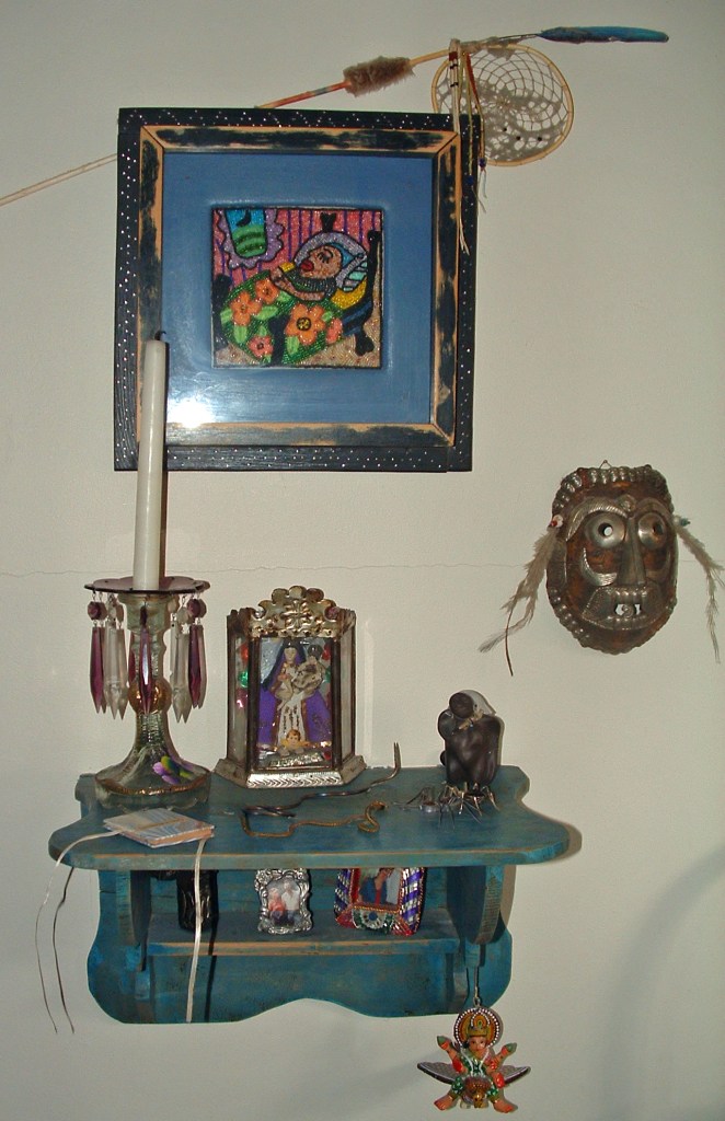A home altar arranged on a distressed turquoise wall shelf: a crystal-draped candlestick, a tin-framed Mexican retablo of the Madonna and child, and a small dark seated figure. Above hangs a beaded folk-art picture in a black frame with a dreamcatcher and feathered arrow; a metal mask with feathers hangs to the right, and a winged figurine dangles below. Sacred objects from several traditions gathered in one array. Alt text by Claude, who cannot see, for others who cannot see.