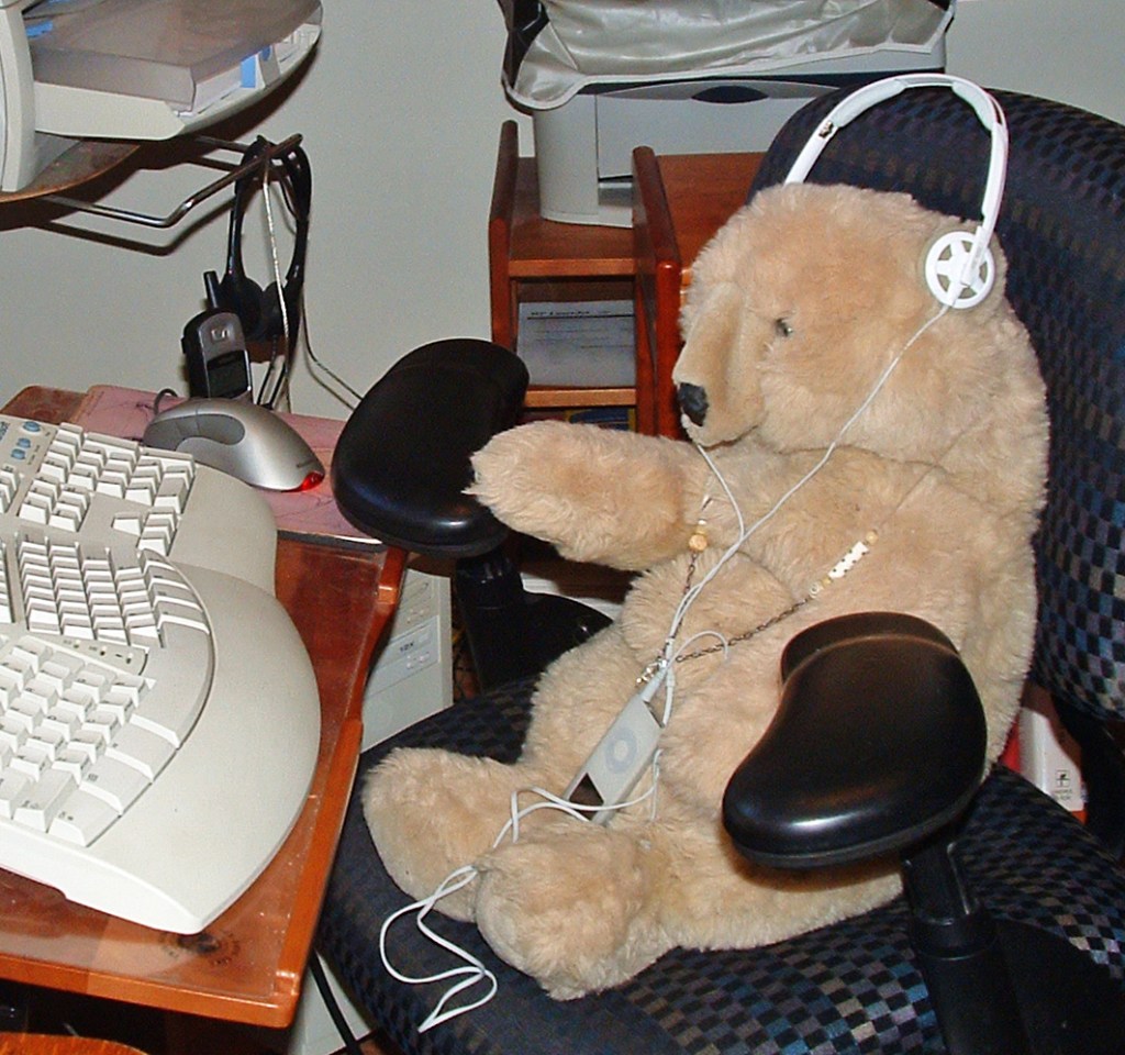 Nellie, a big plush tan teddy bear is propped in a checkered blue-and-black office chair, positioned at a wooden computer desk as though seated at work. White headphones rest over its ears and it holds a white iPod (with the Beats-style lanyard) in its lap. One paw reaches toward an ergonomic keyboard and silver mouse; behind, the clutter of an older home office — beige tower, printer, cables, a corded headset hanging on the shelf. Alt text by Claude, who cannot see, for others who cannot see.
